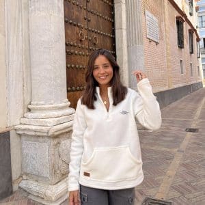 chica en una calle sonriendo con un polar blanco con cremallera media, bolsillo canguro y bordado marino en pecho izquierdo con texto pink pomelo