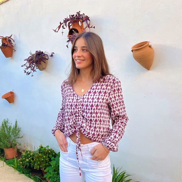 Chica joven con el pelo castaño largo y suelto que viste una blusa manga larga blanca con estampado color berenjena con cordón ajustable en frontal.