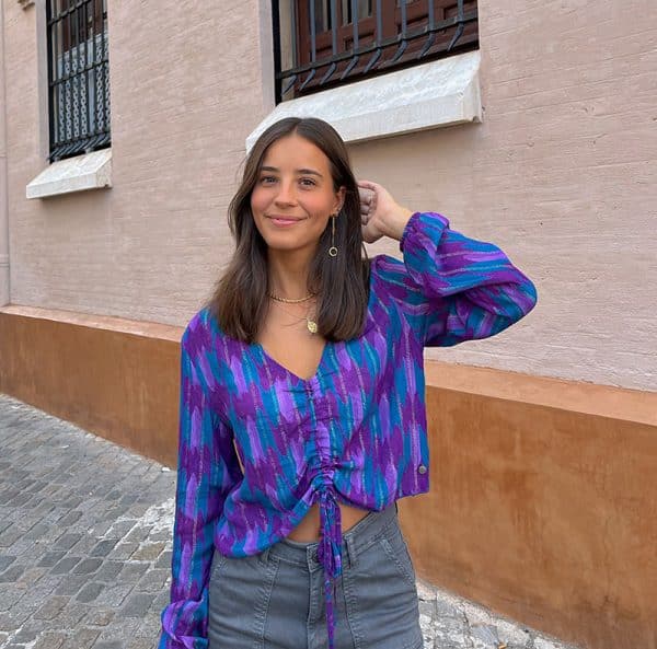 chica en la calle sonriendo con una mano en el pelo con blusa manga larga con goma elástica en puños con cuello pico y cordón ajustable en frontal en color verde azulado con morados e hilo metalizado en plata y dorado