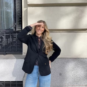 chica en la calle delante de una fachada y ventana con un tejano azul claro y una blusa antracita y blazer de felpa negra