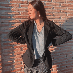 Chica morena de pelo largo mirando hacia el lado derecho que está de pie delante de un muro de ladrillos. Biste una blazer de felpa negra, una camiseta blanca y una falda negra.