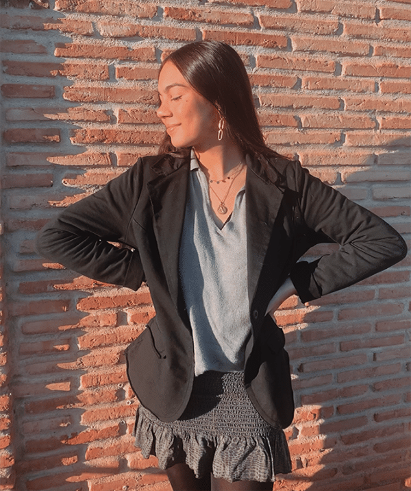 Chica morena de pelo largo mirando hacia el lado derecho que está de pie delante de un muro de ladrillos. Biste una blazer de felpa negra, una camiseta blanca y una falda negra.