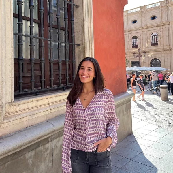 chica posando en la calle con una blusa de escote pico en estampados lila y lúrex y pantalón tejano gris oscuro