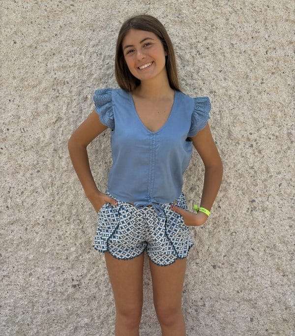 Irene Silva delante de una pared gris con un short estampado azul con fondo blanco con vivos de bolitas azules y una blusa azul con túnel frontal para fruncir cordón y tejido de agujeros en volantes de mangas cortas