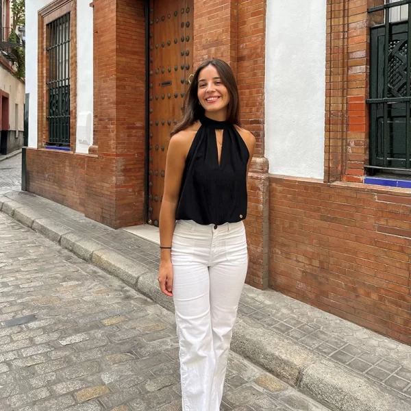 chica posando en la calle con un pantalón blanco con dos bolsillos frontales y blusa cuello halter negra sin mangas con abertura en escote frontal