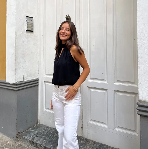 chica posando en la calle con un pantalón blanco con dos bolsillos frontales y blusa cuello halter negra sin mangas con abertura en escote frontal