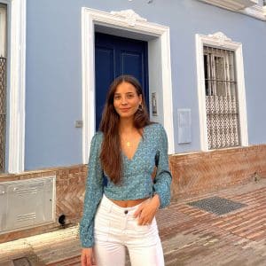 Chica con el pelo largo castaño que está de pie con una mano metida en el bolsillo del pantalón. Viste una blusa estampada corta en tonos azul grisáceo y azul. Fruncida en parte baja de la espalda y unos pantalones blancos.