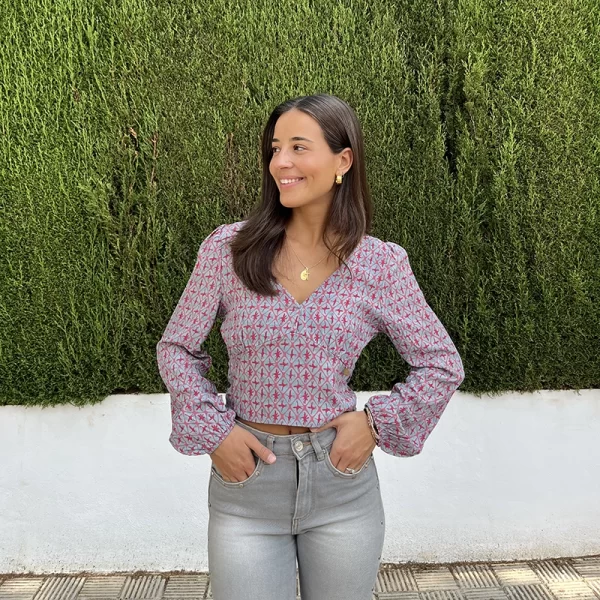 chica en la calle delante de un arbusto verde con blusa modelo Cádiz con estampado verde agua y coral y pantalón gris con tachuelas plateadas