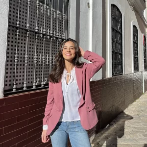 chica posando en la calle con un brazo en el pelo con una blazer de felpa rosa, una blusa blanca de lurex y un pantalón vaquero azul