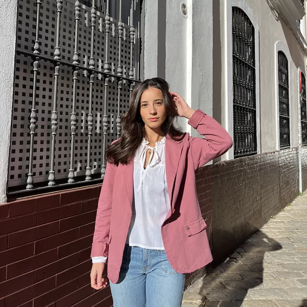 chica posando en la calle con un brazo en el pelo con una blazer de felpa rosa, una blusa blanca de lurex y un pantalón vaquero azul