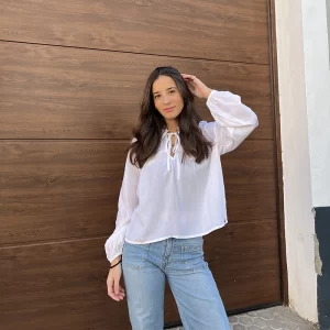 chica con una mano en el pelo delante de una puerta de garaje marrón con un tejano azul con tachuelas en los bolsillos y blusa blanca con lazada en cuello frontal y líneas de hilos de lúrex doradas