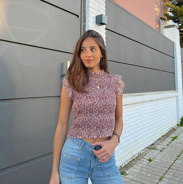 chica delante de una puerta de garaje gris con una blusa fruncida sin mangas en tonos berenjena con lúrex dorado y volantes en mangas y tejano azul con tachuelas en bolsillos frontales y lateral de piernas