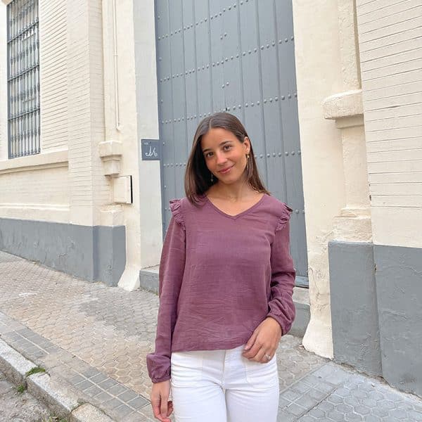 chica delante de una puerta gris con una mano en el bolsillo de su pantalón blanco y una blusa manga larga en color rosa viejo que tiene volantes en hombros