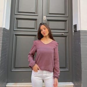 chica delante de una puerta gris con una mano en el bolsillo de su pantalón blanco y una blusa manga larga en color rosa viejo que tiene volantes en hombros