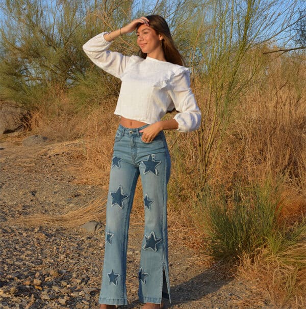 chica en el campo con un pantalón denim con parches de estrellas y blusa blanca manga larga con lúrex plateado