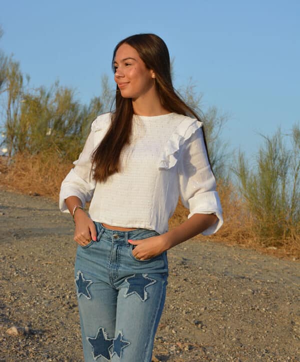 chica en el campo con una blusa blanca con lúrex plata con forro interior blanco y volantes en hombros. Manga larga con goma elástica en puños