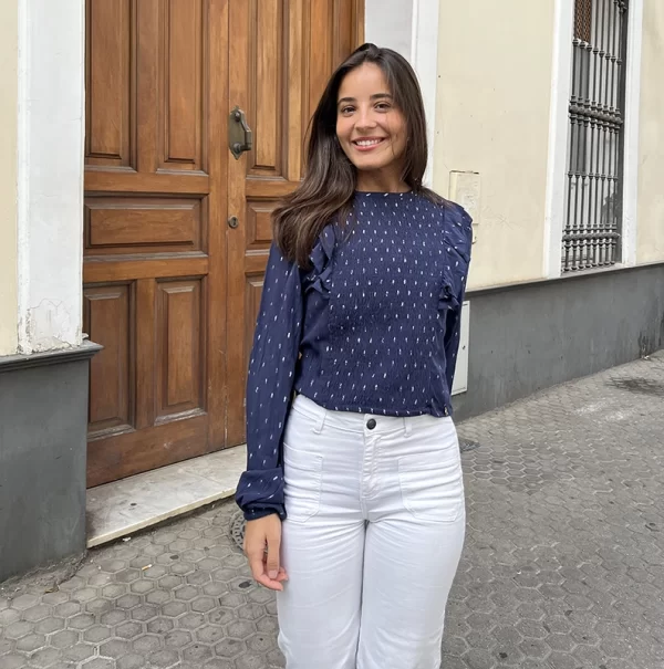 chica en la calle andado de frente con una blusa azul marino con volantes en hombros y detalles con hilo de lúrex plateado. Va con pantalón blanco