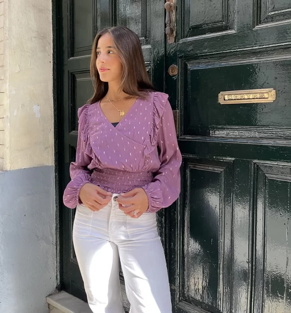 hica posando delante de una puerta verde con una blusa cruzada rosa viejo con lúrex plateado y pantalón blanco