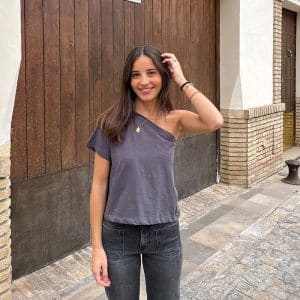 chica sonriendo delante de un portón de madera con una mano en el pelo vestido con un tejano gris oscuro con 2 bolsillos frontales y una camiseta gris antracita asimétrica