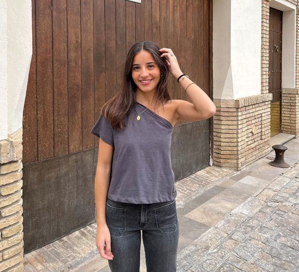 chica sonriendo delante de un portón de madera con una mano en el pelo vestido con un tejano gris oscuro con 2 bolsillos frontales y una camiseta gris antracita asimétrica