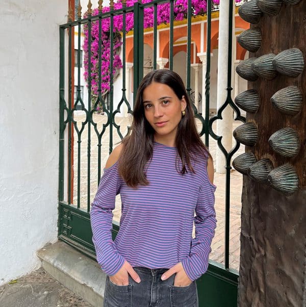 chica con camiseta de rayas azul plomo y rosa con hombros descubiertos y manga larga delante de una reja verde con las manos en los bolsillos del pantalón tejano gris oscuro