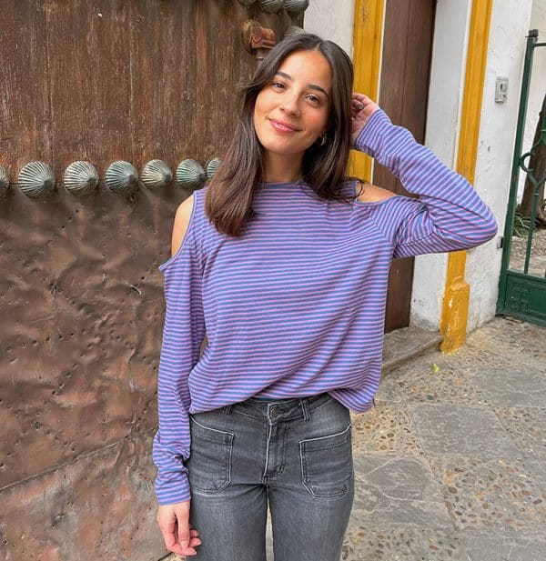 chica con camiseta de rayas azul plomo y rosa con hombros descubiertos y manga larga delante de puerta de madera con las manos en los bolsillos del pantalón tejano gris oscuro