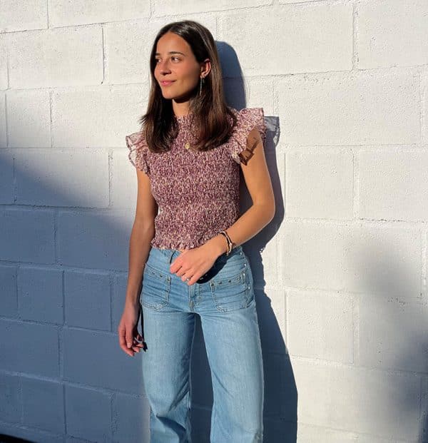 chica delante de una pared blanca con una blusa fruncida sin mangas en tonos berenjena con lúrex dorado y volantes en mangas y tejano azul con tachuelas en bolsillos frontales y lateral de piernas