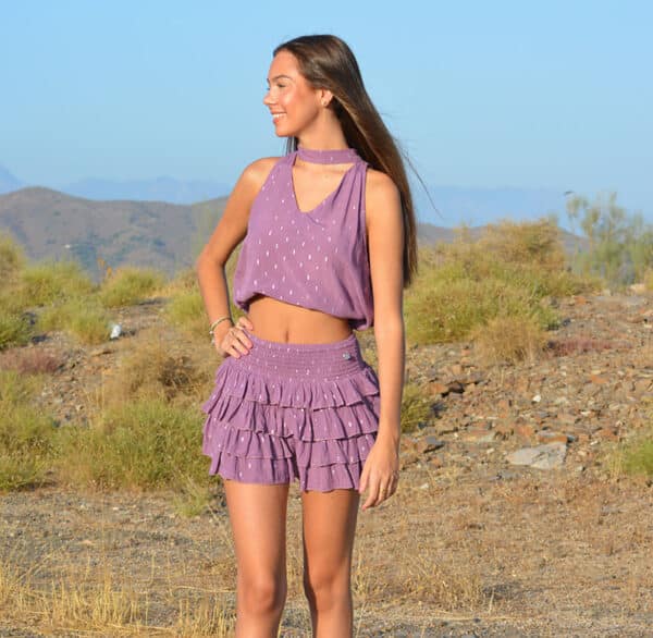 chica en un campo sonriendo con una mano en la cintura vestida con una blusa sin mangas con choker en cuello en rosa con lúrex plata y falda pantalón de 4 volantes en rosa con lúrex plata