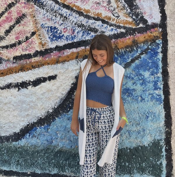 Irene silva delante de una pared llena de grafiti vestido con un pantalón fluido largo en estampado azul y blanco con un top fruncido corto azul y cárdigan blanco sin mangos con bolsillos azules