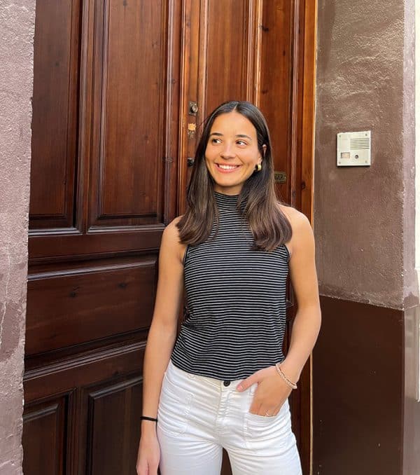 chica sonriendo delante de una puerta de madera con una mano en el bolsillo del pantalón blanco y con un top sin mangas negro de rayas finas blancas y sin mangas