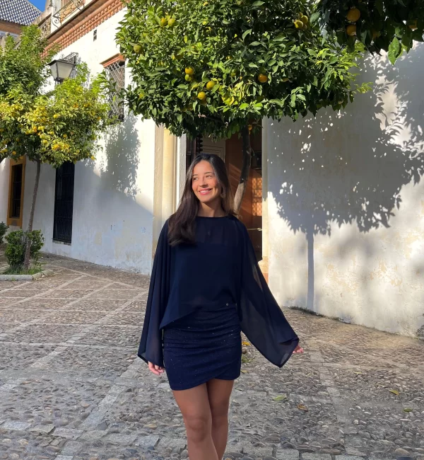 chica sonriendo en un patio con naranjos con un conjunto azul de brillo de falda corta y top banda y una capa de gasa azul