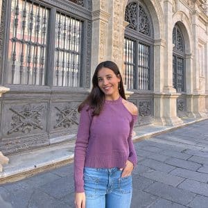 chica en la calle sonriendo con una mano en el bolsillo de su pantalón tejano azul con tachuelas y jersey color berenjena con un hombro descubierto