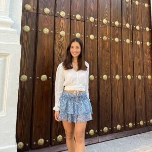 chica delante de portón de madera con una blusa blanca con lúrex plata y falda pantalón de gasa azul estampada con lúrex plata