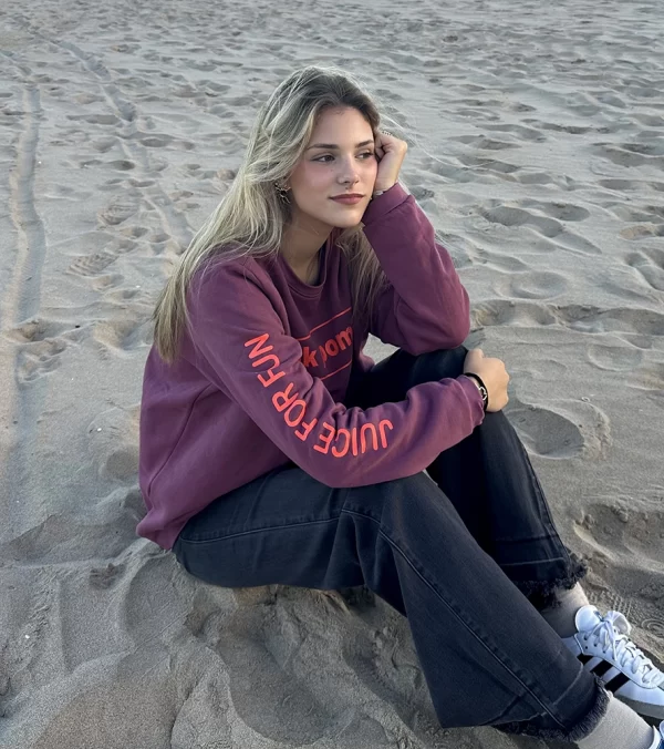 chica sentada en la playa con un pantalón campana gris oscuro y una sudadera de pink pomelo en color burdeos con un estampado grande en espalda y manga en coral flúor