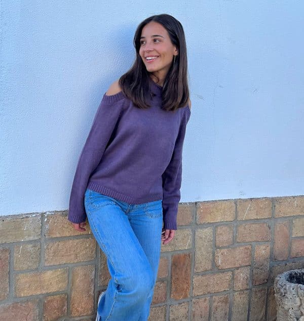 chica sonriendo y mirando a un lateral apoyada en una pared blanca con ladrillos en el bajo con un jersey morado de hombros descubiertos y pantalón campana azul con tachuelas en frontal y lateral