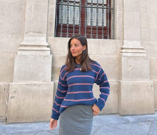 chica de frente mirando a un lado con un jersey azul de rayas rosas con hombros descubiertos y falda larga gris