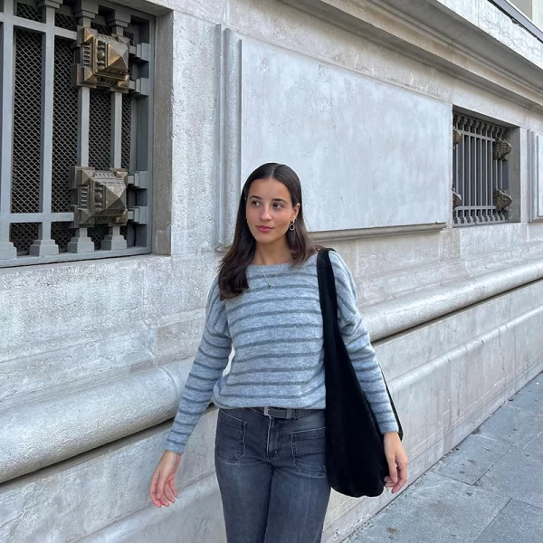 chica andando por la calle con un bolso negro en el hombro vestida con un jersey de rayas grises y pantalón gris oscuro