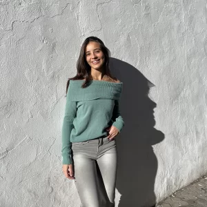 chica apoyada en una pared con un jersey verde de hombros descubiertos con la vuelta dada y pantalón gris claro con tachuelas