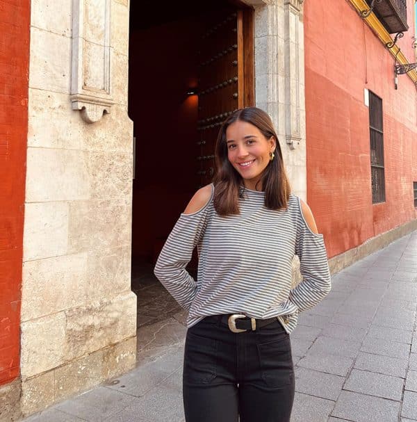 chica en la calle con las manos en la espalda con una camiseta de rayas antracita rosa claro con hombros descubiertos y pantalón negro