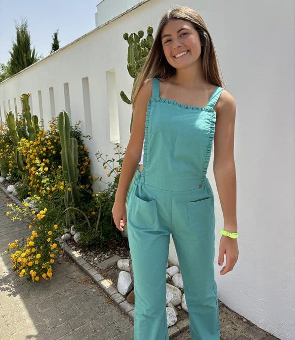 Irene silva en el exterior de una casa moderna con pared blanca y flores sonriendo con un mono verde con bolsillos frontales y volantito discreto en la pieza frontal