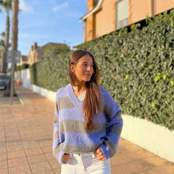 chica con manos en los bolsillos del pantalón sonriendo con un jersey oversize de rayas azul lila con gris y pantalón blanco