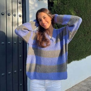 chica con los brazos levantados y manos detrás de la cabeza sonriendo con un jersey oversize de rayas azul lila con gris y pantalón blanco