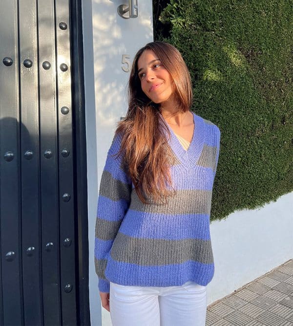 chica con los brazos atrás sonriendo con un jersey oversize de rayas azul lila con gris y pantalón blanco