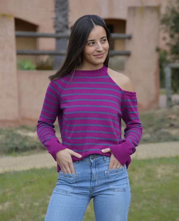 chica con las manos en los bolsillos del pantalón azul tejano con un jersey tricotosa de color fucsia orquídea con rayas en color azul y con hombro izquierdo descubierto. Detalle de chapa metálica Pink Pomelo en el bajo lateral izquierdo.