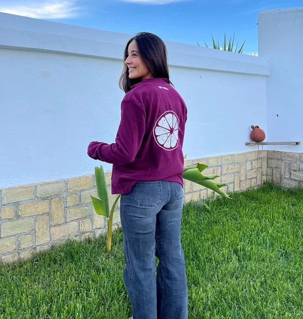 chica en un jardín de espaldas con un polar berenjena con bordado blanco en espalda de una rodaja grande de un pomelo y bordado pequeño en cuello con el nombre de la marca pink pomelo y con un pantalón tejano azul