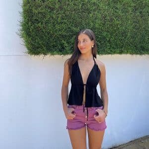 chica posando en la calle con un short denim rosa desflecado en bajo y cierre con botones plateados y un top negro anudado al cuello y escote pico