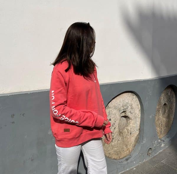 chica delante de una pared blanca y gris con una sudadera coral con estampado blanco en pecho izquierdo, brazo derecho y espalda