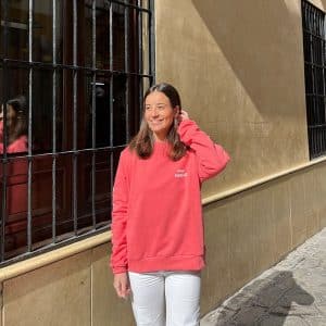 chica delante de una ventana de rejas con una sudadera coral con estampado blanco en pecho izquierdo, brazo dereho y espalda