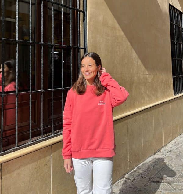 chica delante de una ventana de rejas con una sudadera coral con estampado blanco en pecho izquierdo, brazo dereho y espalda