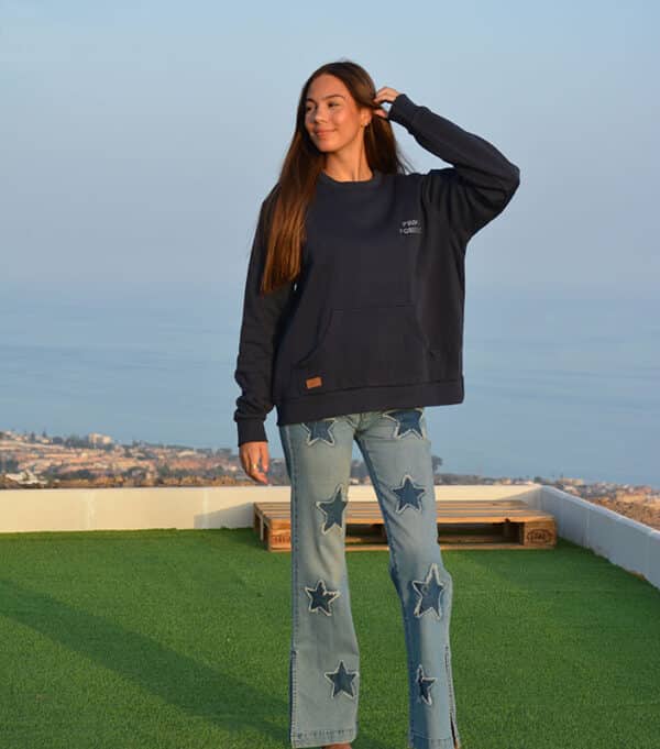 chica con una mano en el pelo con un tejano azul con parches de estrellas y sudadera azul marino con texto pink pomelo en pecho izquierdo hecha de strass y bolsillo canguro